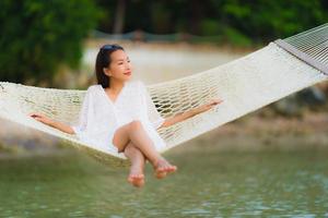 stående vacker ung asiatisk kvinna som sitter på hängmattan runt havsstranden havet för att koppla av foto