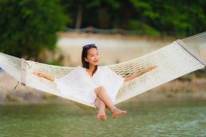 stående vacker ung asiatisk kvinna som sitter på hängmattan runt havsstranden havet för att koppla av foto