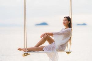 stående vacker ung asiatisk kvinna som sitter på gungan runt stranden havet för avkoppling foto