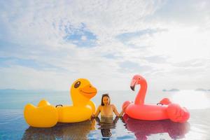 stående ung asiatisk kvinna på uppblåsbar flottör gul anka och rosa flamingo runt utomhuspool i hotell och semesterort foto