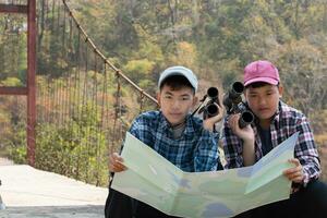 asiatisk Pojkar innehar kikare och lokal- nationell parkera Karta, vilar och läsning information i Karta nära sling bro av lokal- nationell parkera under natur och sommar läger av dem, tonåren' aktivitet begrepp. foto