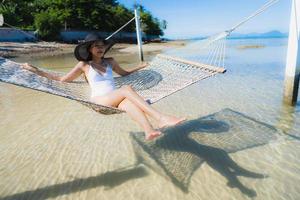 stående vacker ung asiatisk kvinna som sitter på hängmattan runt havsstranden havet för att koppla av foto