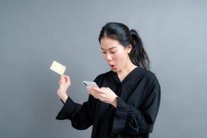 ung asiatisk tjej som visar plastkreditkort medan du håller mobiltelefonen
