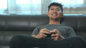 asiatisk ung man har roligt spelar video spel sitta soffa på Hem. foto