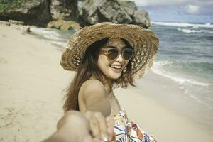 en Lycklig ung asiatisk kvinna är leende till de kamera och pekande på de strand i gunungkidul, Indonesien. foto