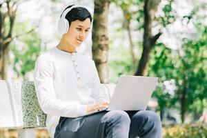 stilig asiatisk mansammanträde med bärbar dator för att arbeta i parken foto