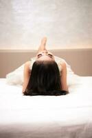 ett asiatisk kvinna i en vit klänning är liggande på en vit säng med henne svart hår lösa i en hotell foto