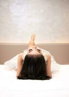 ett asiatisk kvinna i en vit klänning är liggande på en vit säng med henne svart hår lösa i en hotell foto