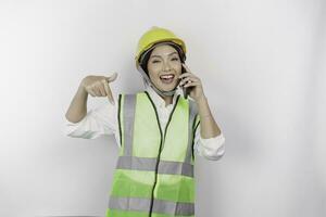en leende asiatisk kvinna arbetskraft bär säkerhet hjälm och väst, talande på henne telefon och pekande till kopia Plats Nedan henne, isolerat förbi vit bakgrund. arbetskraftens dag begrepp. foto