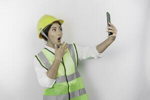 en porträtt av en chockade asiatisk kvinna arbetskraft bär säkerhet hjälm och väst medan innehav henne telefon med henne mun bred öppna, isolerat förbi vit bakgrund. arbetskraftens dag begrepp foto