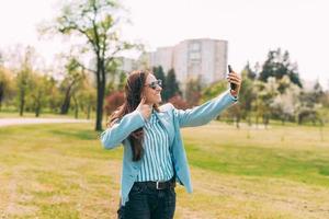 glad ung kvinna i blå kostym som går i park och gör selfie med smarttelefonen och visar upp tummen utomhus foto