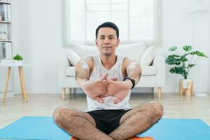 uppkopplad klass Träning. asiatisk mogna man stretching i sportkläder framställning stretching för ben på matta yoga i levande rum interiör och leende. Lycklig sportsman träna förbi honom slef. sport på Hem. foto