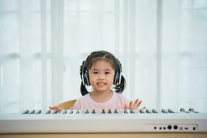 asiatisk söt flicka leende och bär vit hörlurar spelar inlärning uppkopplad piano musik i de levande rum på Hem. de aning av aktiviteter för de barn på Hem under karantän. musik inlärning studie. foto
