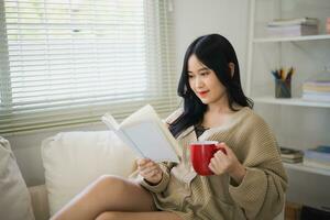 glad asiatisk kvinna läsning bok och dryck varm kaffe medan Sammanträde på soffa soffa i levande rum på Hem. asiatisk kvinnor relex läsning bok leende på soffa i de hus. aktivitet hobby på hus begrepp. foto