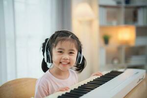 asiatisk söt flicka leende och bär vit hörlurar spelar inlärning uppkopplad piano musik i de levande rum på Hem. de aning av aktiviteter för de barn på Hem under karantän. musik inlärning studie. foto