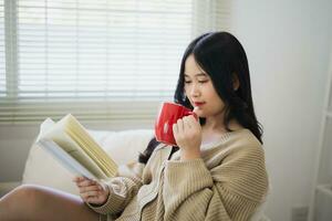 glad asiatisk kvinna läsning bok och dryck varm kaffe medan Sammanträde på soffa soffa i levande rum på Hem. asiatisk kvinnor relex läsning bok leende på soffa i de hus. aktivitet hobby på hus begrepp. foto