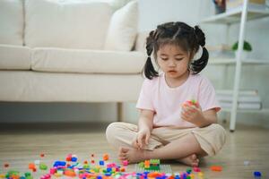 glad asiatisk flicka Lycklig och leende spelar färgrik LEGO leksaker, Sammanträde på de levande rum golv, kreativt spelar med lego, byggnad färgrik strukturer kreativitet tänka. inlärning utbildning. foto