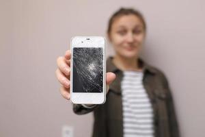 frustrerad ung kvinna med mobiltelefon med trasigt skärmglas. telefonskärmen måste repareras. isolerad på grå bakgrund. selektivt fokus på smartphone foto