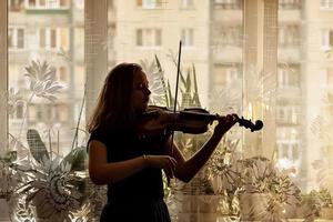 silhuett av en ung flicka, en musiker. spelar fiol i fönstrets bakgrund foto