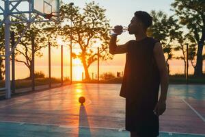 svart man håller på med sporter i morgon, dricka vatten på basketboll domstol på soluppgång foto