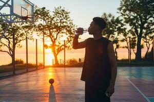 svart man håller på med sporter i morgon, dricka vatten på basketboll domstol på soluppgång foto