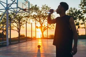 Häftigt svart man håller på med sporter i morgon, dricka vatten på basketboll domstol på soluppgång foto