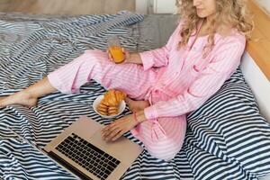söt ung Söt blond kvinna i rosa pyjamas Sammanträde på säng arbetssätt på bärbar dator, frilansare på Hem foto