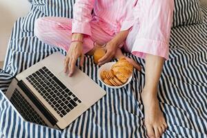 söt ung Söt blond kvinna i rosa pyjamas Sammanträde på säng arbetssätt på bärbar dator, frilansare på Hem foto