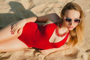 ung skön blond kvinna solbad på sand strand i röd simning kostym, solglasögon foto