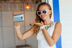 attraktiv kvinna i vit klänning i sommar Kafé i solglasögon använder sig av telefon tar Foto