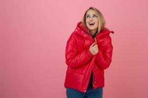 blond Lycklig attraktiv aktiva kvinna Framställ på rosa bakgrund i färgrik vinter- ner jacka av röd Färg, har roligt, värma täcka mode trend, leende foto