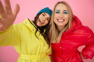 två attraktiv flicka vänner kvinnor tar selfie Foto på rosa bakgrund i färgrik vinter- jacka av ljus röd och gul Färg har roligt tillsammans, värma täcka sportkläder mode trend, galen rolig