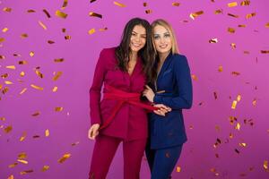 två attraktiv kvinnor fira ny år på violett bakgrund i eleganta färgrik kväll kostymer av lila och blå Färg, vänner har roligt tillsammans, mode trend, gyllene konfetti fest humör foto