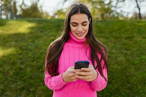 ung leende kvinna i rosa Tröja gående i grön parkera använder sig av telefon foto