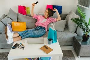 Lycklig leende kvinna i rosa skjorta på soffa på Hem handla uppkopplad foto