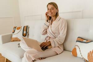 Söt leende kvinna arbetssätt på bärbar dator på Hem på säng i morgon- i pyjamas foto
