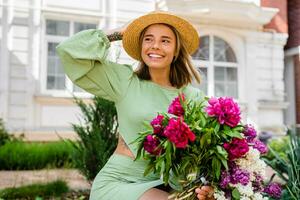 skön ung kvinna i sommar stil utrusta leende Lycklig gående med blommor i stad gata foto
