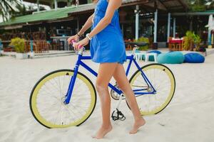ung attraktiv leende kvinna i blå klänning gående på tropisk strand med cykel foto
