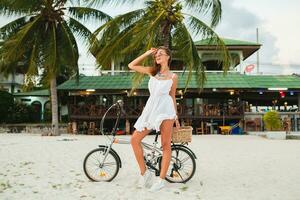 ung attraktiv leende kvinna i vit klänning ridning på tropisk strand på cykel foto
