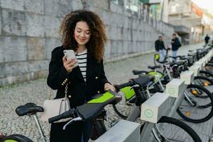 Söt lockigt kvinna hyra en cykel i gata med ett app foto