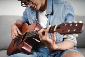 njut av stilig asiatisk man som tränar eller spelar gitarr i soffan i vardagsrummet foto