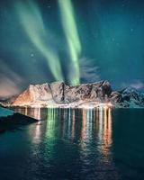 aurora borealis, norrsken över snöiga berg med glödande by i hamnoy på lofoten öar