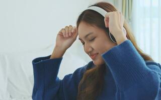 Lycklig asiatisk kvinna dans och lyssnande till musik med hörlurar på säng. foto
