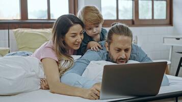 caucasian familj av tre använder sig av bärbar dator medan liggande på säng tillsammans, bläddring internet eller tittar på film. foto