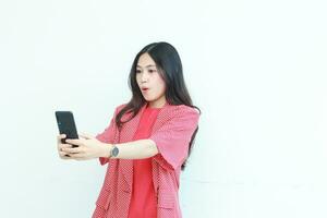 porträtt av skön asiatisk kvinna bär röd utrusta ser på mobil telefon med överraskad uttryck foto