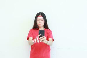 porträtt av skön asiatisk kvinna bär röd utrusta fira indonesien oberoende dag innehav mobil telefon med tänkande uttryck foto