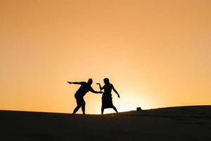 silhuetter av en lycklig ung par kille och flicka på en bakgrund av orange solnedgång i sandöknen foto