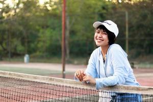 vacker asiatisk kvinna med kort hår, hatt och ler bredt på tennisbanan foto