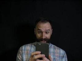 snygg man med skägg och mustasch ser in i en mobiltelefon med ett förvånat ansikte på en svart bakgrund