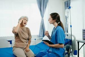 läkare i vit kostym ta anteckningar medan diskuterar och asiatisk äldre, kvinna patient vem liggande på säng med tar emot salin lösning i sjukhus eller klinik. foto